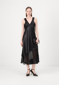 Black sleeveless asymmetrical dress with a satin finish and layered sheer fabric, paired with black pointed-toe heels and a small black handbag.