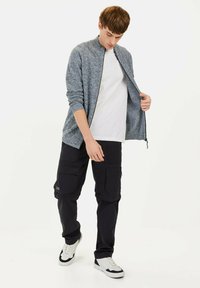 Leichte, strukturierte dunkelblaue Jacke mit Reißverschluss, über einem weißen T-Shirt getragen. Schwarze Cargo-Hosen mit Seitentaschen. Weiße Turnschuhe mit schwarzen Akzenten.