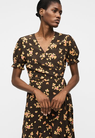 Γυναίκα που φοράει σκούρο φόρεμα τύπου wrap με φουσκωτά κοντά μανίκια και κίτρινο και ροζ λουλουδάτο σχέδιο, με τα χέρια ελαφρώς σταυρωμένα στη μέση.