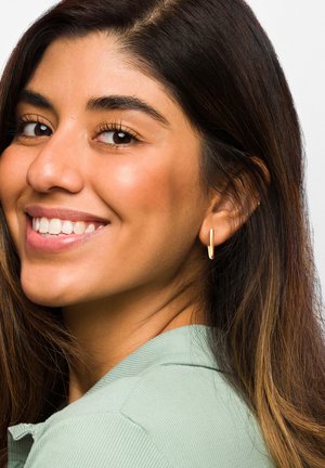 Goldene Creole mit einem eleganten, verlängerten Design. Das Model trägt ein mintgrünes Hemd mit Kragen, während lange dunkle Haare über ihre Schulter fallen.