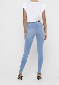 Vaqueros ajustados de un azul claro con una cintura de media altura, que presentan un diseño de cinco bolsillos y costuras sutiles. Combinados con una blusa blanca de manga corta.