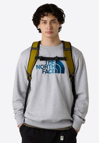 Gray sweatshirt with blue logo; model wears a yellow backpack with black straps; textured fabric; relaxed fit; casual design.
