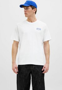 Camisa de algodón blanca con mangas cortas y un texto gráfico azul "ORIGINALS SINCE 1990" en el lado izquierdo del pecho. Pantalones de mezclilla oscuros. Gorra azul.