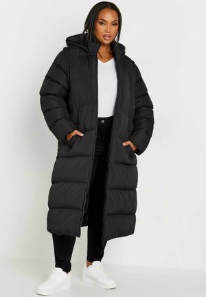Abrigo puffer largo negro con capucha, que presenta un cierre de cremallera, bolsillos laterales y un patrón acolchado. Combinado con pantalones negros y zapatillas blancas.