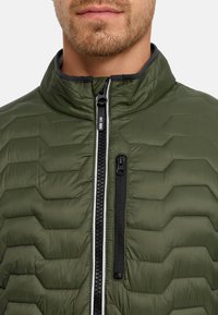 Skæggede mand iført grøn quiltet jakke med sort lynlås og brystlomme-lynlås, synlig fra nakke til øvre bryst.