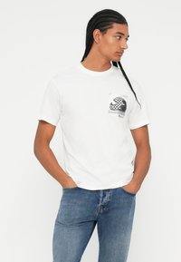 Λευκή βαμβακερή μπλούζα με κοντά μανίκια, που διαθέτει σχέδιο μαύρο-άσπρο καρό στην τσέπη του στήθους. Έχει στρογγυλή λαιμόκοψη και χαλαρή γραμμή.