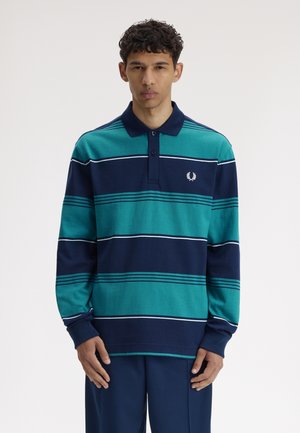 Jeune homme portant un polo à manches longues rayé bleu sarcelle et bleu marine avec col, debout devant un fond clair uni.