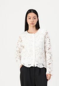 Cardigan bianco in pizzo con motivo floreale, maniche lunghe, scollatura rotonda e chiusura frontale con bottoni; indossato sopra pantaloni neri.