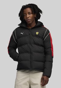 Zwarte pufferjack met rode accenten en witte piping, voorzien van een Puma-logo en Ferrari-embleem. Ritssluiting aan de voorkant en verstelbare capuchon.
