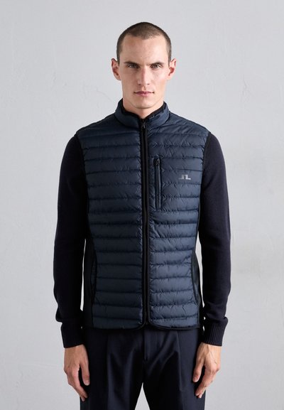 Gilet imbottito blu con colletto alto, design trapuntato orizzontale, zip frontale e tasca sul petto sinistro; abbinato a un maglione scuro sottostante.