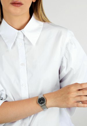Silberne Armbanduhr aus Edelstahl mit schwarzem Zifferblatt, silbernen Stundenmarkierungen und einem schlanken, minimalistischen Design, getragen an einem Handgelenk mit hellem Hautton.