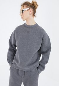 Grauer Pullover aus weichem Material mit Rundhalsausschnitt und fallenden Schultern. Verfügt über ein kleines Prägungslogo auf der Brust.
