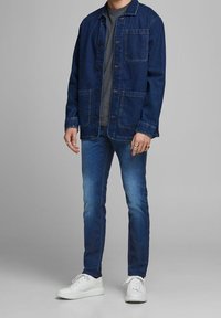 Mörkblå denimjacka med två framfickor, lager-på-lager över en grå skjorta. Bärs med slim-fit blå jeans och vita sneakers.