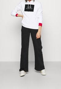 Felpa bianca con polsini rossi, caratterizzata da un logo rettangolare nero; abbinata a pantaloni neri a gamba larga e sneakers bianche.