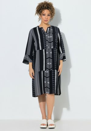 Frau mit lockigem Haar, die ein knielanges Kleid mit schwarz-grauem geometrischem Muster, Dreiviertelärmeln und weißen offenen Schuhen trägt.