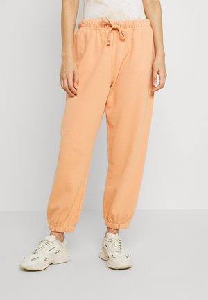 Perzikkleurige joggingsbroek met een elastische tailleband en trekkoord, met cuffs bij de enkels en een zachte textuur. Draagt het met lichtgekleurde sneakers.