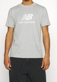 Hombre con una camiseta gris clara de New Balance y pantalones negros, de pie con los brazos relajados frente a un fondo blanco liso.