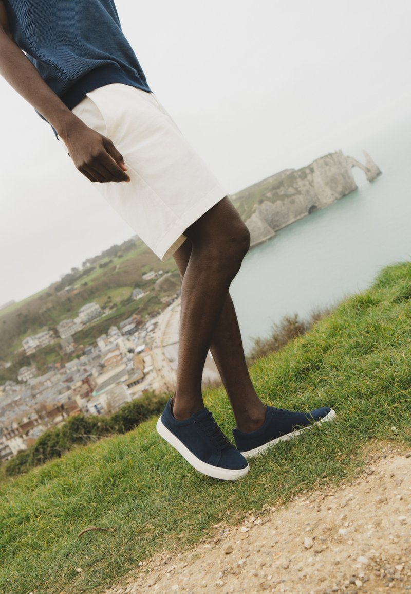 Personne portant un short blanc, des baskets bleu marine et un t-shirt bleu marine, debout sur une falaise herbeuse surplombant une ville côtière et une arche marine.