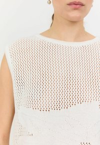 Lättviktig, vit stickad topp med en lös nätmönster, rund halsringning och subtil textur med ett lövmönster i nederkant.
