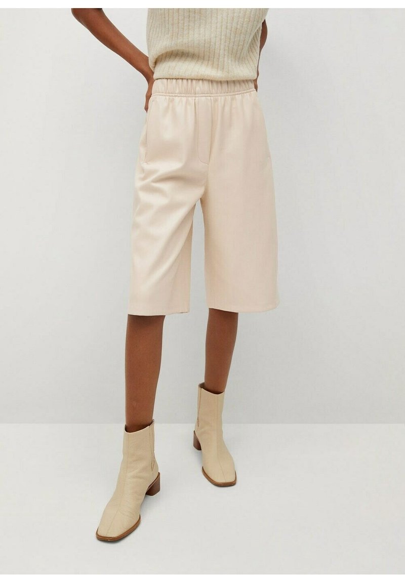 Femme portant un haut sans manches en maille crème, un short ample beige longueur genou et des bottines beiges, debout contre un mur uni.