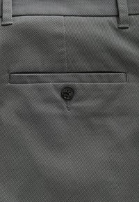 Pantalones de tela gris con un patrón de textura diagonal, un bolsillo trasero con un botón negro y presillas para cinturón estándar.