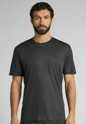 Mann mit dunklem lockigem Haar und Bart, der ein schlichtes dunkelgraues kurzärmeliges T-Shirt mit Brusttasche trägt, steht vor einem hellgrauen Hintergrund.