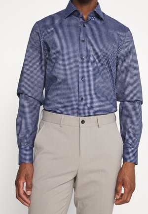 Camisa de vestir a cuadros en azul marino y blanco, con cuello abotonado, mangas largas, combinada con pantalones beige. Botones negros, corte estructurado.