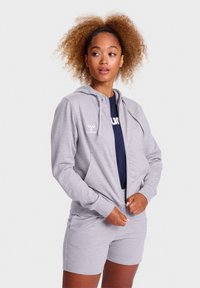 Šedá mikina na zips s kapucňou na sťahovanie, prednými vreckami a detailom loga, kombinovaná so šedými šortkami, na modeli na pozadí s jednofarebným pozadím.
