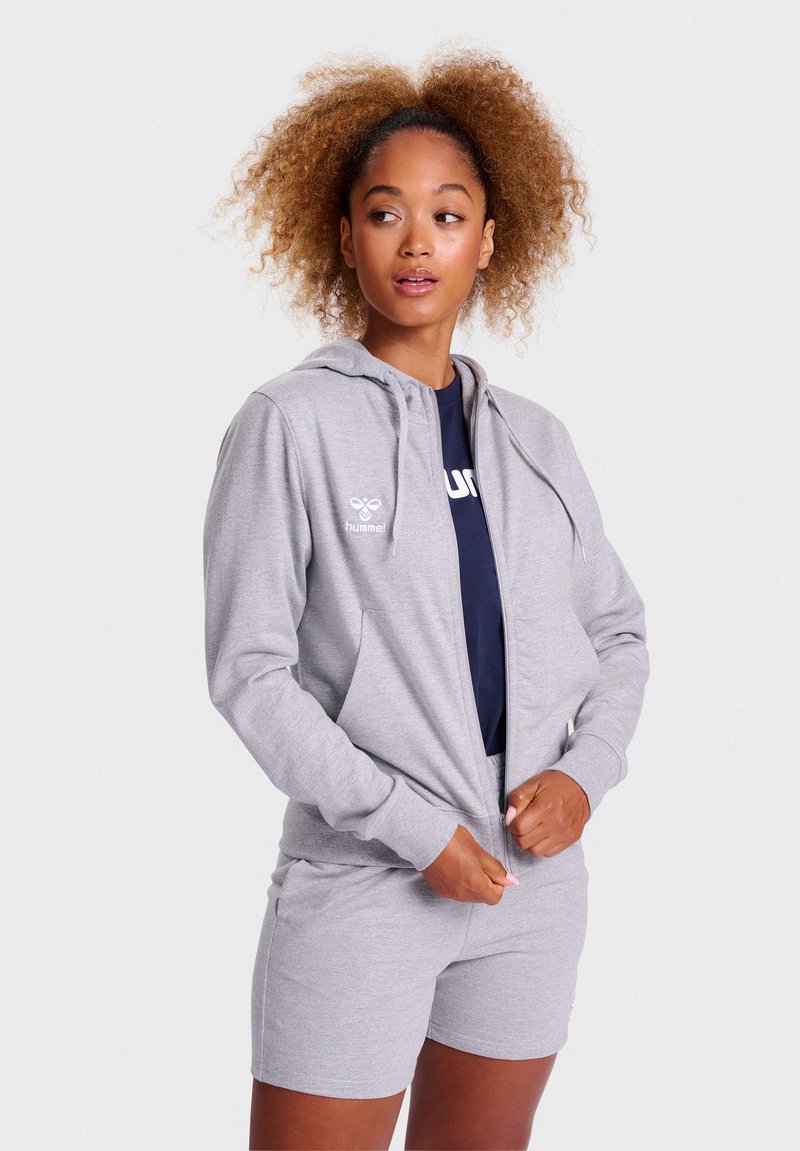Šedá mikina na zips s kapucňou na sťahovanie, prednými vreckami a detailom loga, kombinovaná so šedými šortkami, na modeli na pozadí s jednofarebným pozadím.