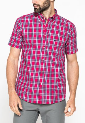 Uomo con una camicia a maniche corte a quadri rossi e blu e pantaloni grigi, che guarda a sinistra con le braccia rilassate.