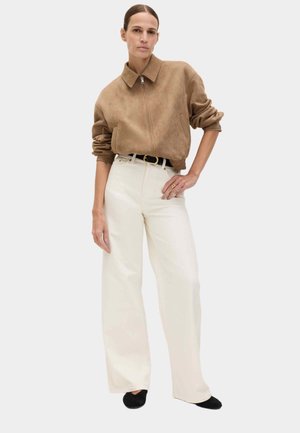 Mujer de pie con una chaqueta con cremallera color tostado, pantalones blancos de pierna ancha y cintura alta, cinturón negro y zapatos negros, con la mano en la cadera.