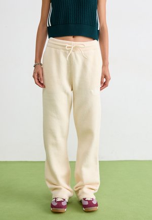 Osoba ubrana w kremowe spodnie dresowe ze sznurkiem, ciemnozielony krótki sweter oraz fioletowo-białe trampki stojąca na zielonej podłodze.