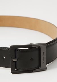 Svart lærbelt med en jevn tekstur og en matt svart metallspenne, med "Calvin Klein"-logoen gravert på metalldetaljene.