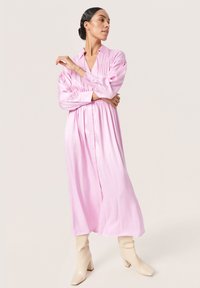 Abito a maniche lunghe, con bottoni, in satin rosa chiaro, cinture in vita con delicate pieghe; indossato con stivali alla caviglia beige con tacco alto.