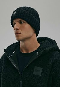 Czarna, prążkowana czapka beanie z naszywką logo, noszona z ciemnym polarowym kapturem z zamkiem błyskawicznym i teksturowanym materiałem.