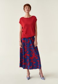 Rood kortarmig topje gecombineerd met een blauwe maxirok met patroon en rode abstracte vormen, aangevuld met lichtblauwe high-heeled schoenen.