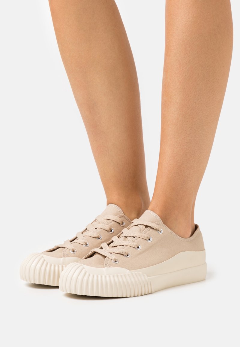 Beige canvas sneakers med rundade, räfflade gummisulor och fem snörningsöglor. Minimalistisk design med en slät yta och lätt konstruktion.