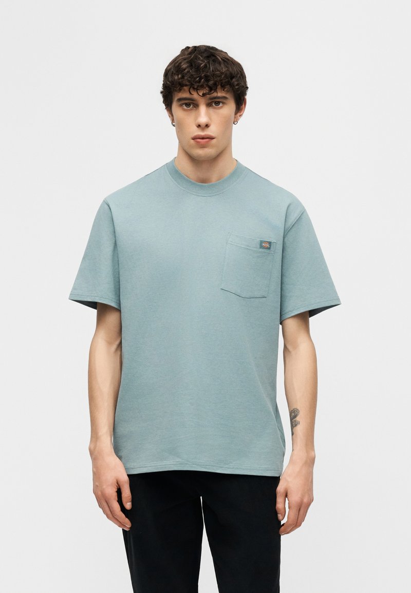 Giovane uomo con capelli ricci che indossa una maglietta azzurra con tasca e pantaloni neri, in piedi davanti a uno sfondo chiaro uniforme.