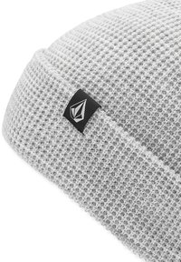 Bonnet gris avec un motif de tricot texturé, arborant une étiquette noire avec un logo blanc sur le côté. Forme arrondie et matériau flexible.