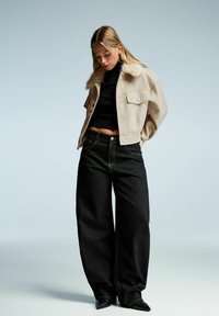 Beige wolmix jas met nep bontkraag, zwarte geribbelde top en donkere wijde jeans. Transparante textuur, ontspannen pasvorm en minimale details.