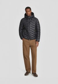 Giubbotto imbottito nero con motivo a cucitura orizzontale, tasche con zip e cappuccio, abbinato a pantaloni marroni e scarpe scure. Design semplice e moderno.