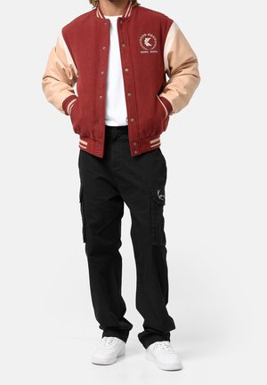 Giacca varsity in lana rossa con maniche in pelle beige, polsini e colletto a costine. Maglietta bianca, pantaloni cargo neri e sneakers bianche.