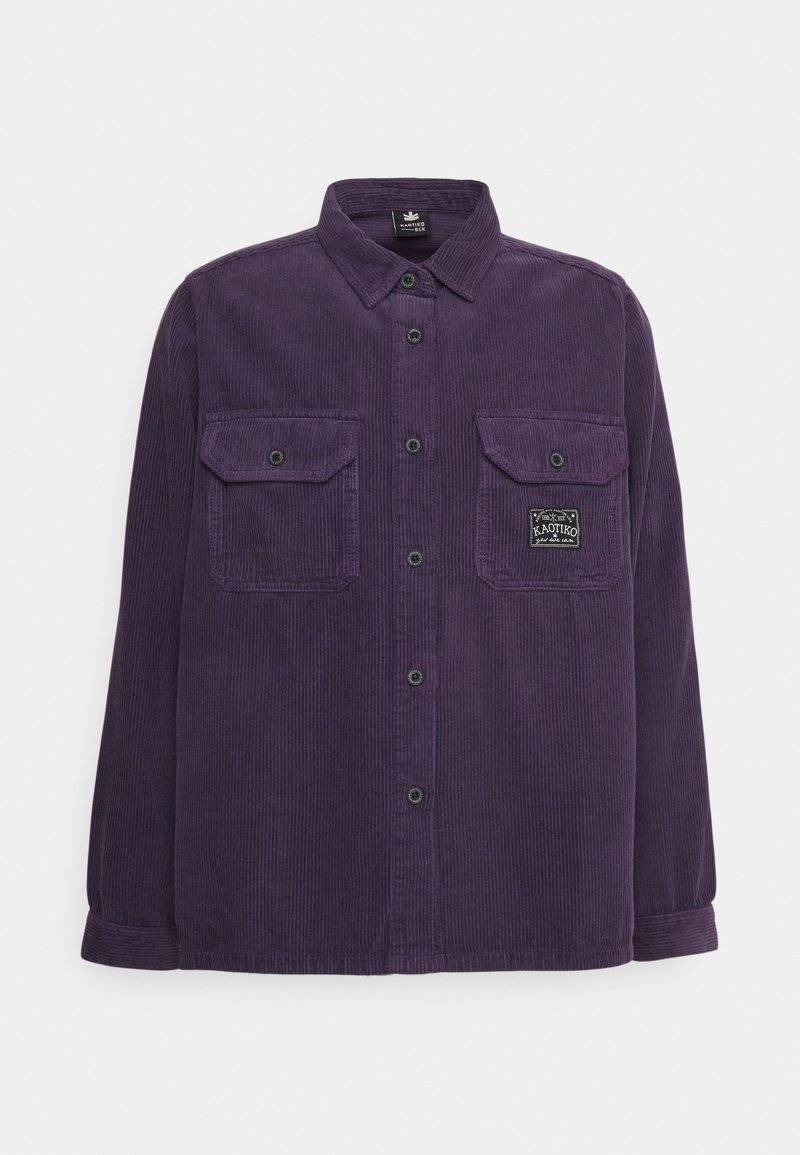 Camisa de pana morada con cierre de botones en la parte delantera, dos bolsillos en el pecho y una superficie texturizada. Presenta detalles en negro en los botones y un parche de logo.
