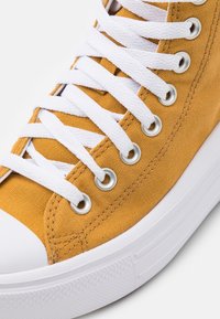 Zapatilla alta de lona amarilla con cordones blancos, ojales plateados y una puntera de goma blanca. Tejido texturizado y detalle de costura diagonal.