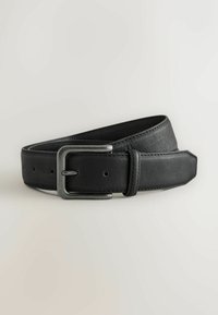 Ceinture en cuir noir avec une boucle rectangulaire argentée mate, roulée et posée sur un fond clair neutre.