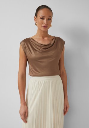 Femme portant un haut marron en satin sans manches avec un col drapé et une jupe plissée blanche taille haute, debout devant un fond uni.