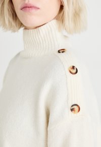 Suéter de punto color crema con un cuello alto acanalado y cuatro botones decorativos marrones en el hombro izquierdo. Textura suave, diseño casual.