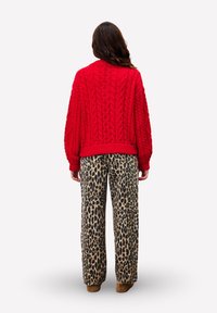 Une personne portant un pull en maille rouge et un pantalon à impressions léopard, se tenant de dos contre un fond uni.