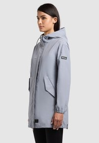 Naine kannab helehalli kapuutsiga mantlit küljetaskute ja elastsete varrukaservadega, seisab külje pealt tavalise tausta ees.