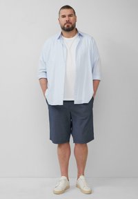 Jasnoniebieska koszula zapinana na guziki na białym t-shircie, w zestawie z ciemnymi szortami i białymi trampkami, stojąca na neutralnym tle.